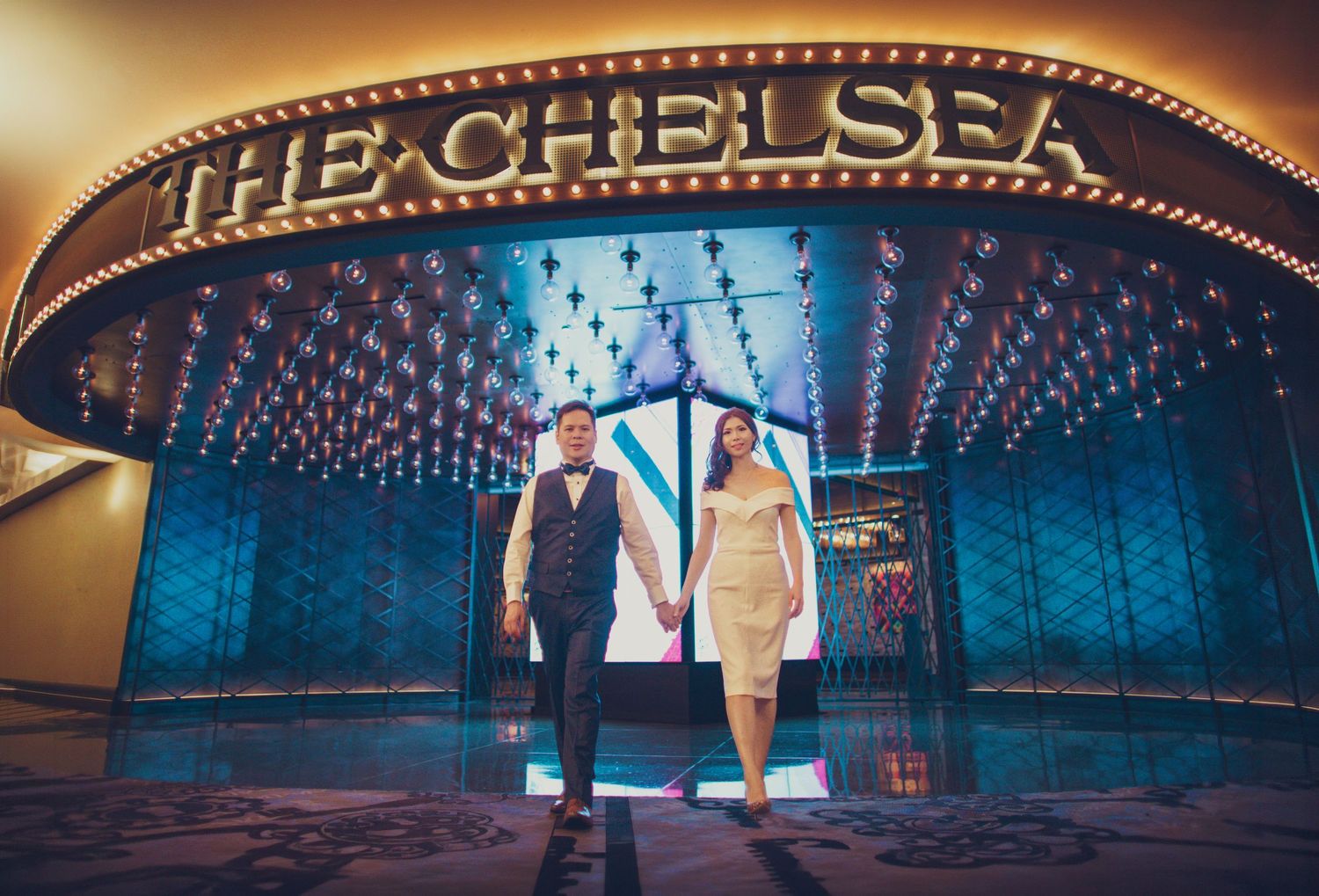 Elegant couple walking hand in hand in front of The Chelsea venue marquee lights.