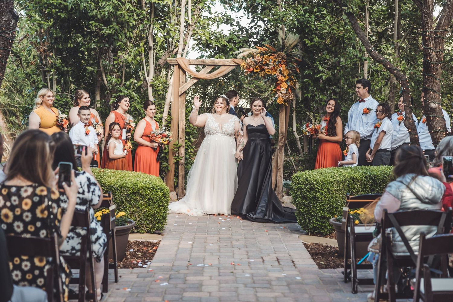 Two brides in white and black gowns walk down aisle at outdoor garden wedding.