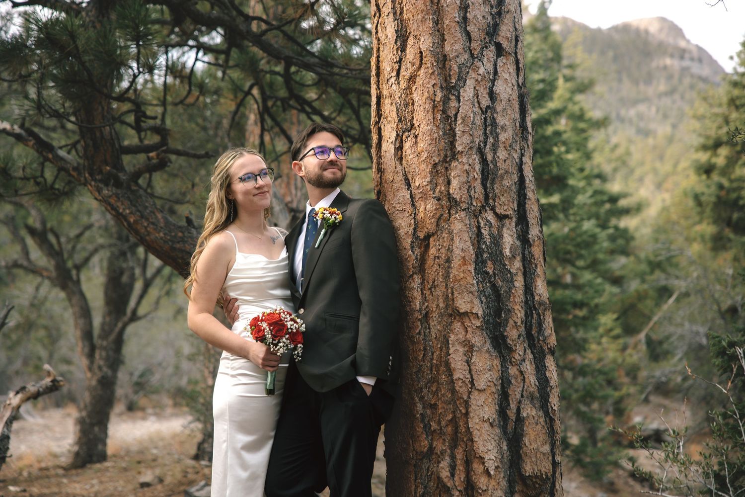 Bride and groom pose beside a tall pine tree in a mountain forest wedding setting.