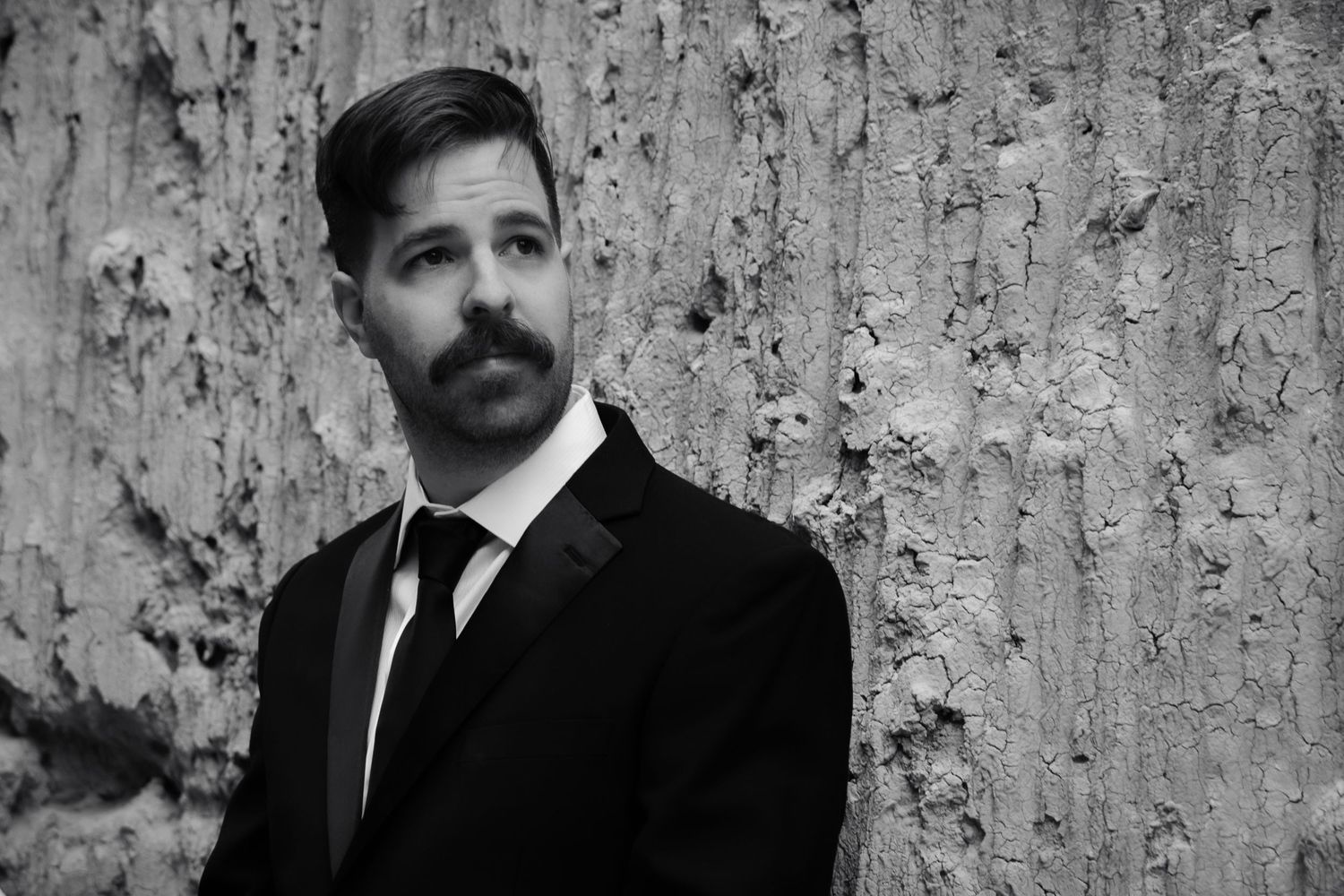 Black and white portrait of a groom in a suit with a mustache leaning against a textured stone wall.