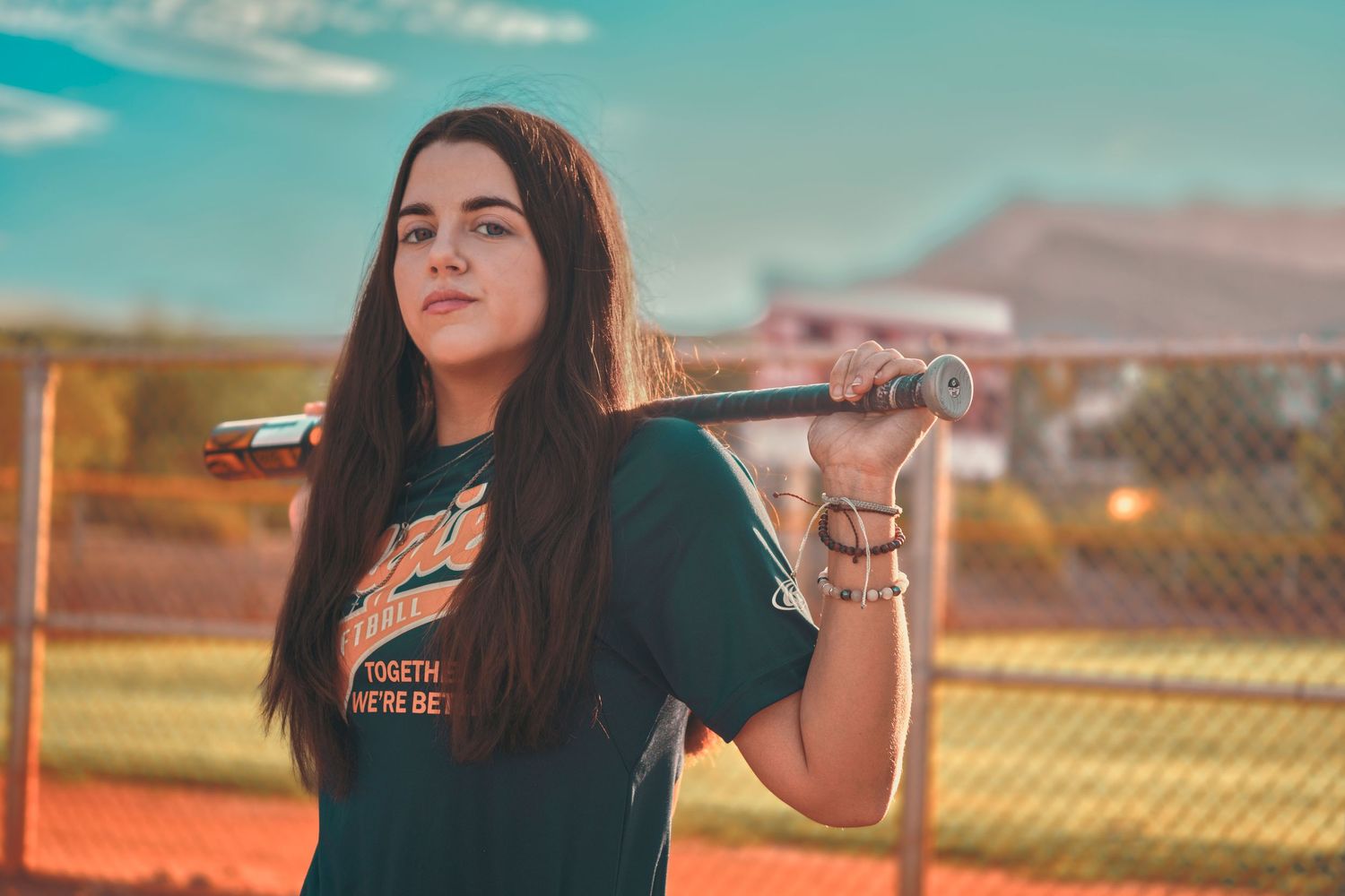 Young female softball player holding a bat over her shoulder on a sports field, wearing a green team t-shirt.