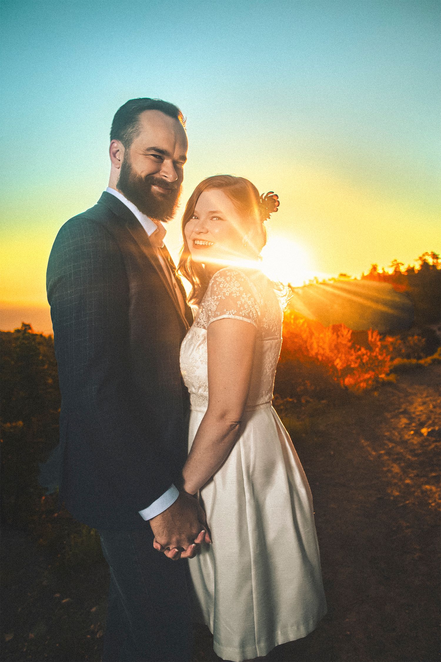 Couple holding hands at sunset, groom in dark suit, bride in white lace dress, golden sunburst behind them outdoors.