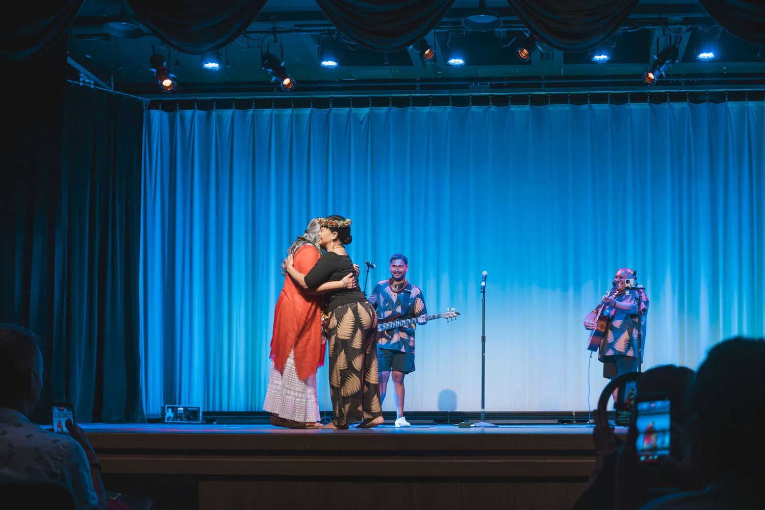 Performers in colorful costumes on a blue-lit stage with musicians playing guitar during a cultural show.