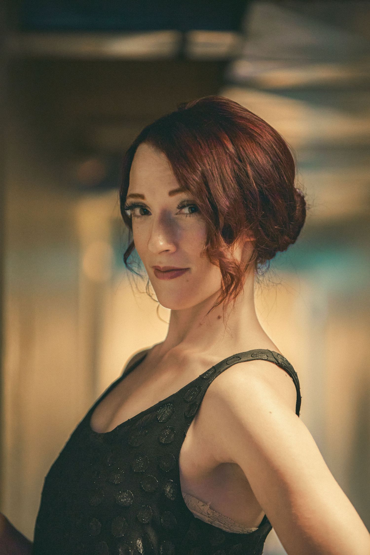 A woman with auburn updo hair wearing a black lace dress poses elegantly in warm ambient lighting.