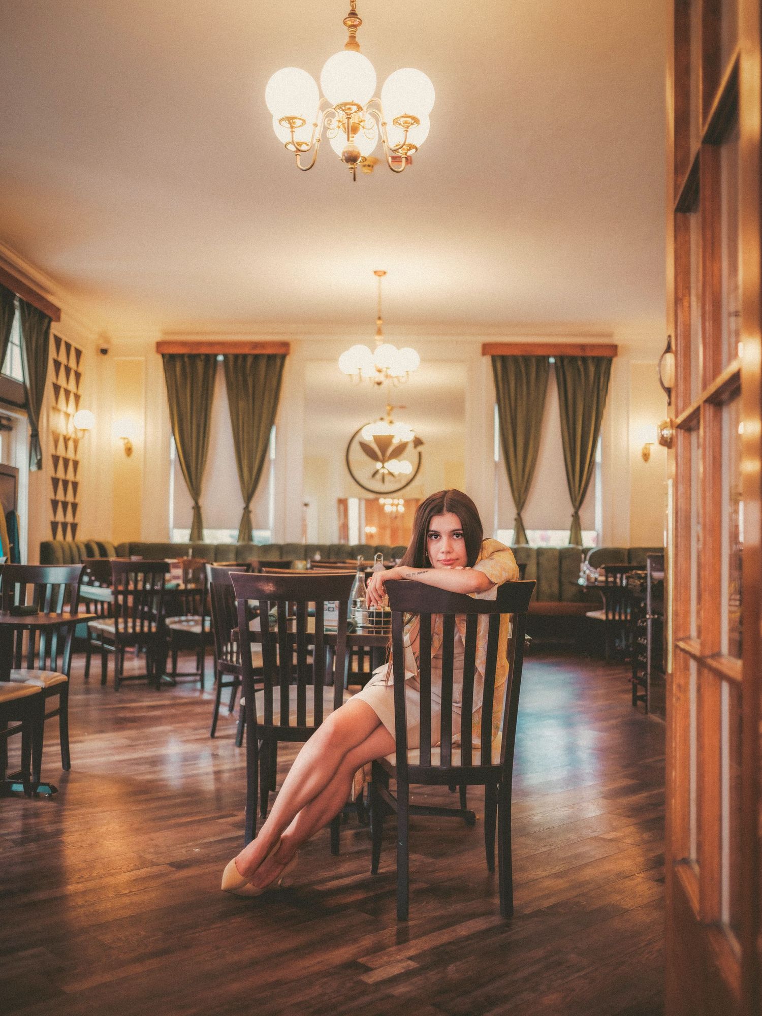 Young woman posing elegantly on a chair in a vintage restaurant with chandeliers and hardwood floors.