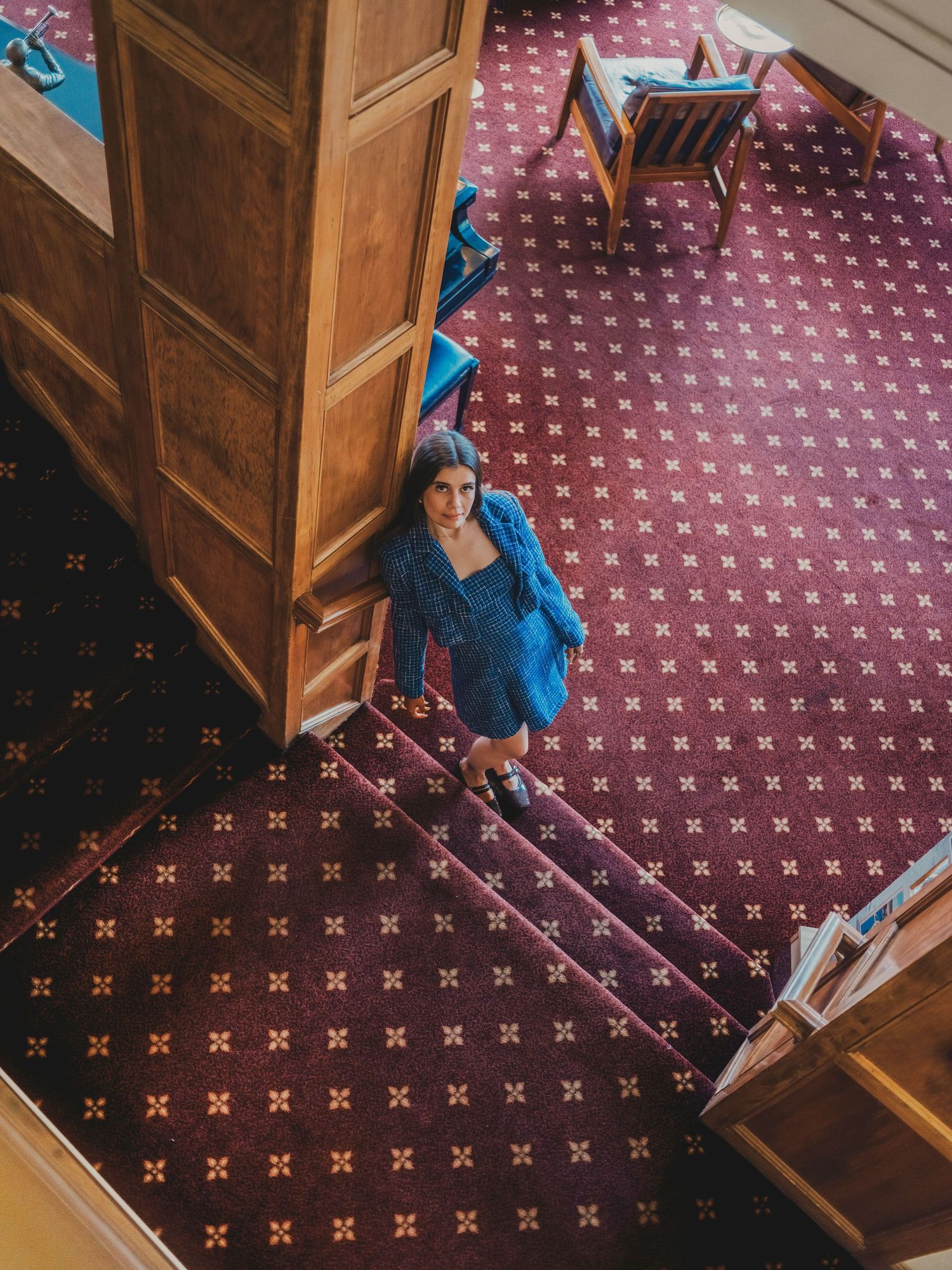 Aerial view of a woman in a blue dress standing on patterned red carpet near wooden doors in an elegant venue.