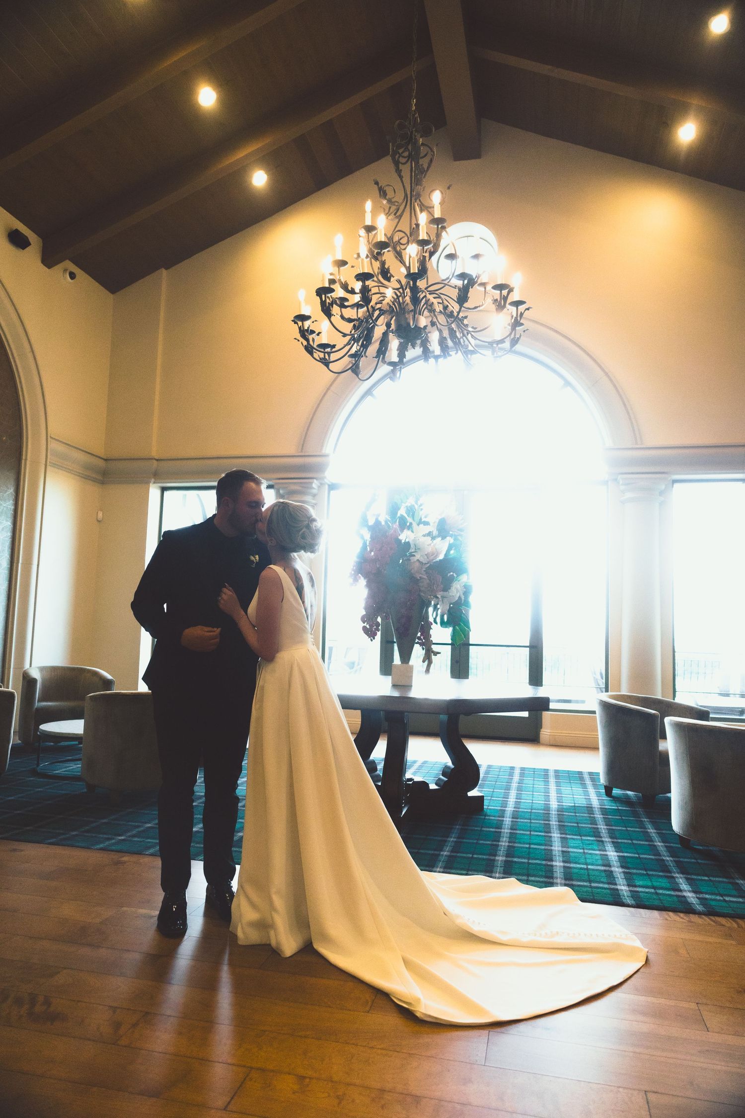 A bride and groom share a romantic kiss in a grand room with a chandelier and arched window.