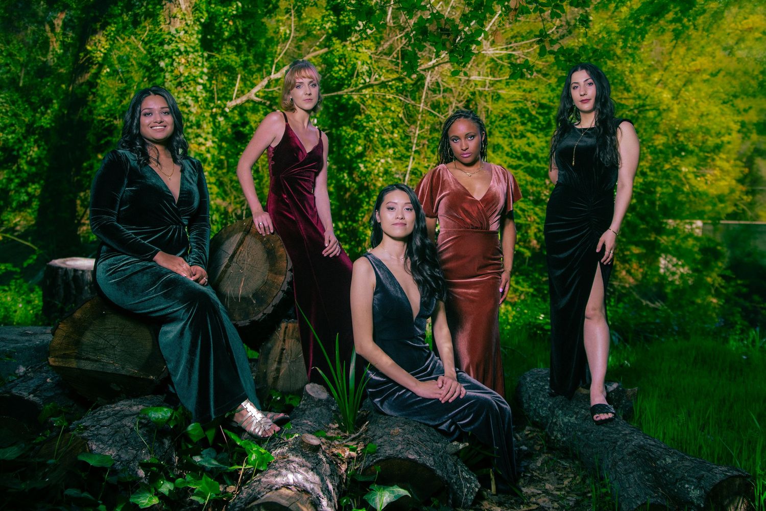 Five women in elegant velvet gowns pose on rocks in a lush green outdoor setting.