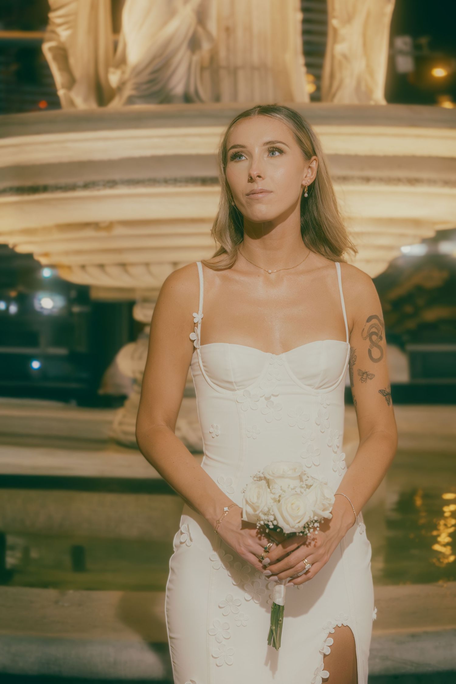 Elegant bride in white spaghetti strap gown holds white rose bouquet before illuminated fountain at night.