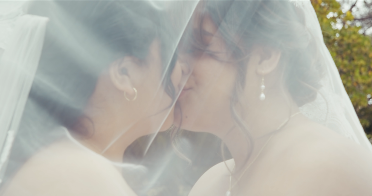 Two brides sharing a kiss under a flowing white veil outdoors.