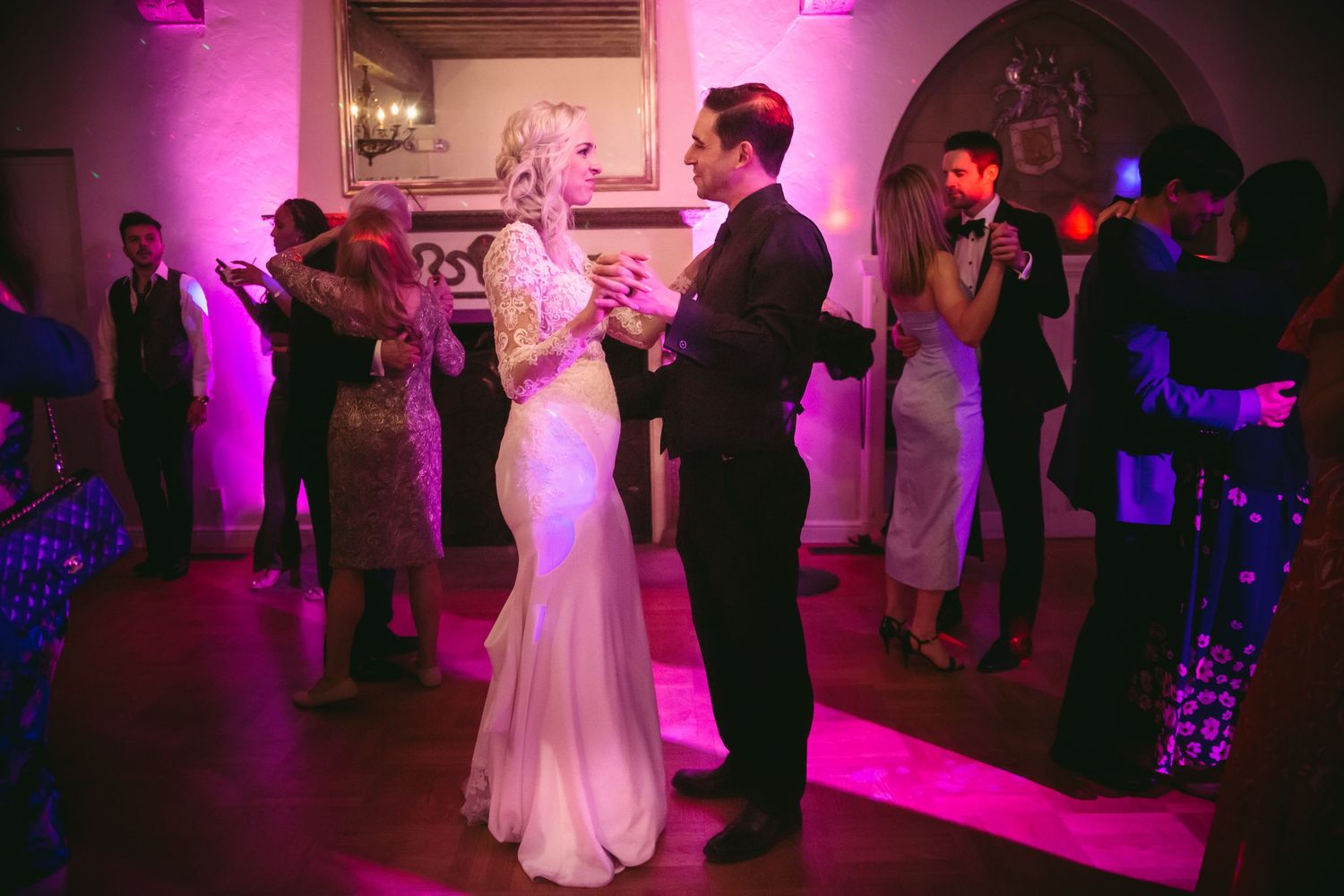 Bride and groom share their first dance surrounded by guests in a romantic pink-lit ballroom.
