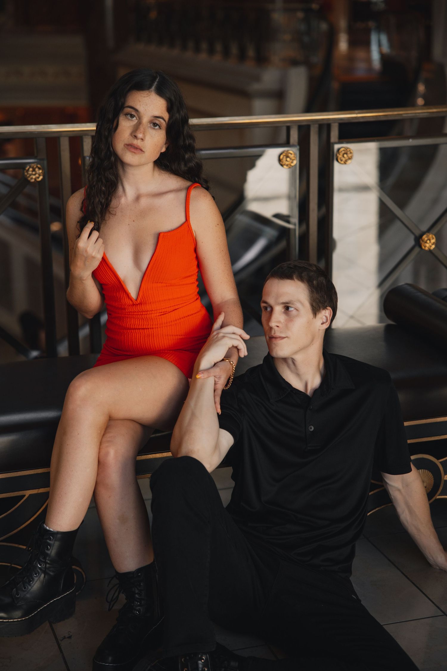 Couple posing elegantly indoors, woman in red bodysuit sitting on railing, man in black holding her hand.