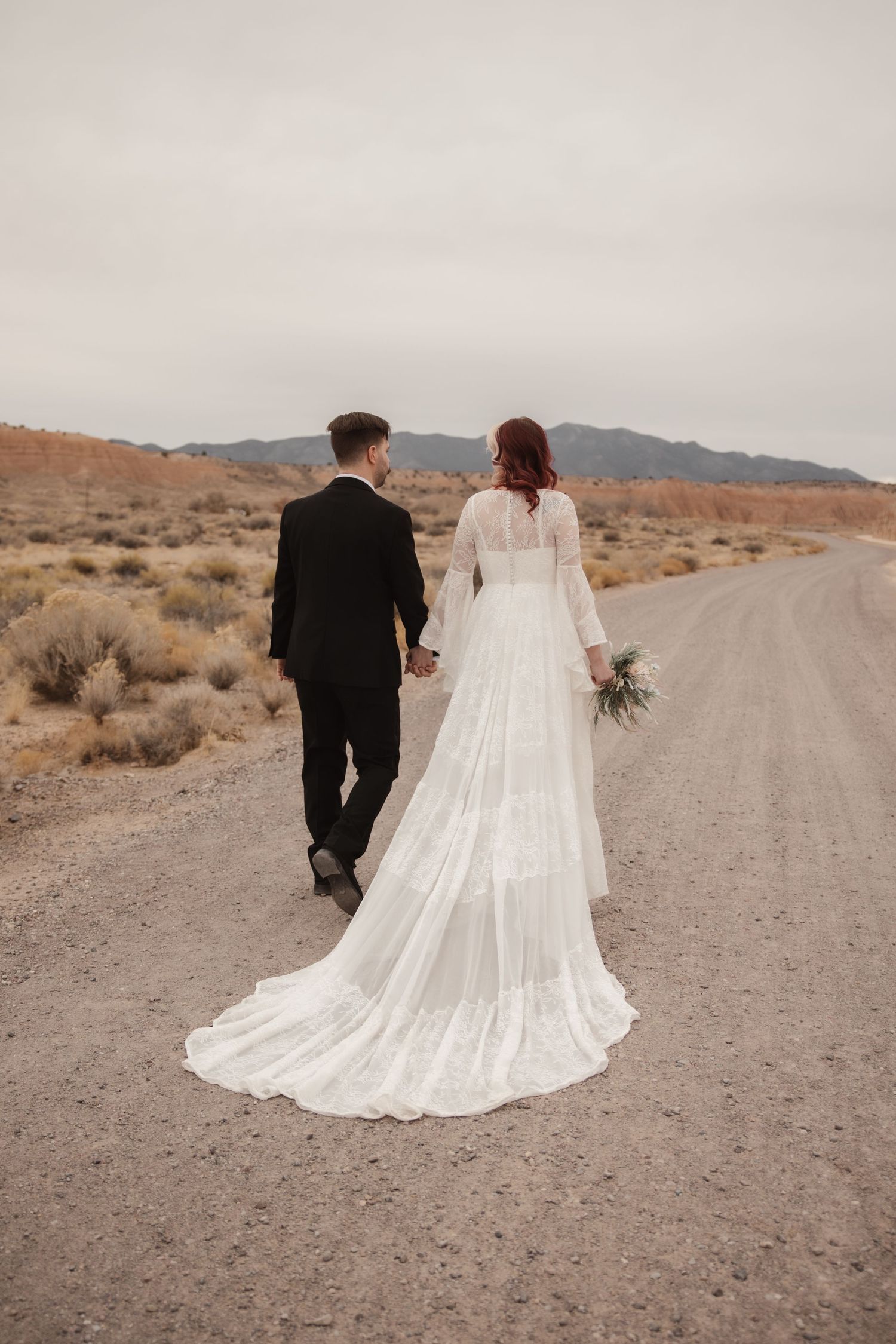 Bride in long-sleeved lace gown with train and groom walk down desert road holding hands.