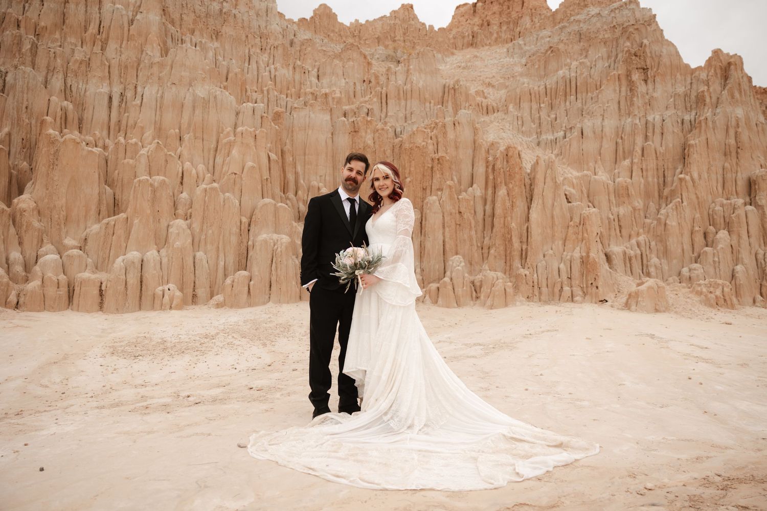 Bride and groom embrace in desert setting with towering red sandstone rock formations in the background.