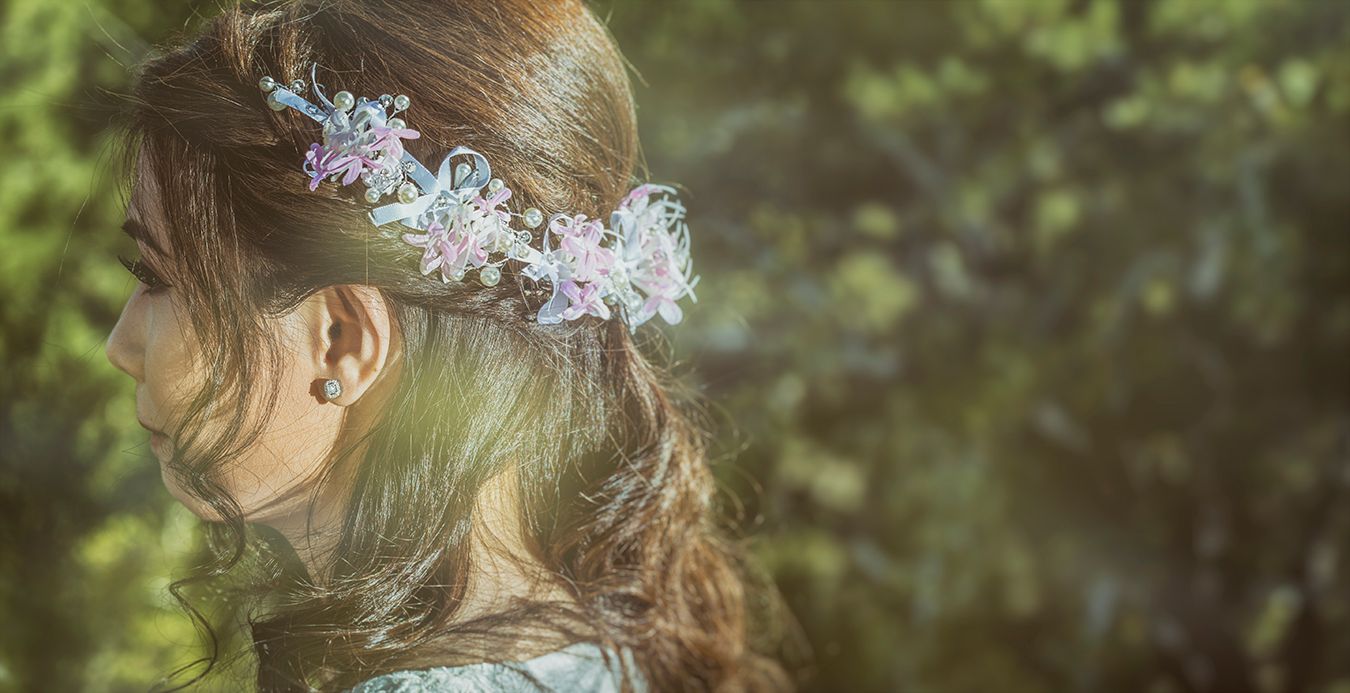 Young woman with elegant floral hair accessories in pink and blue, photographed in a lush green garden setting.