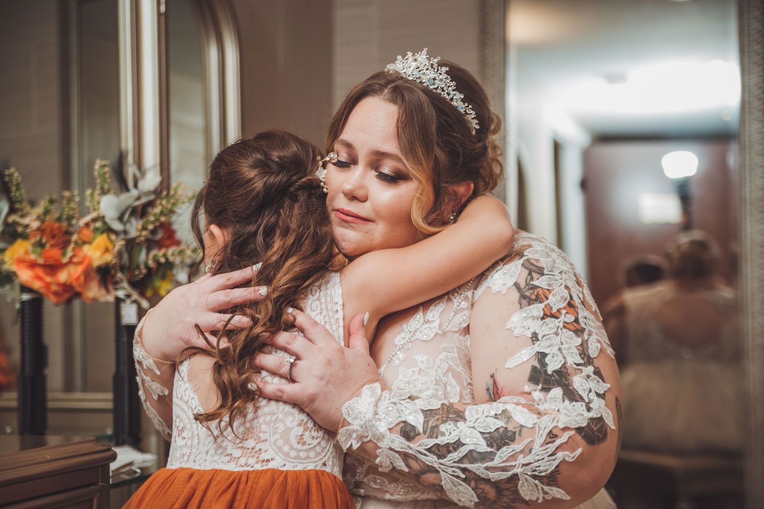 Bride in lace wedding dress shares emotional hug with bridesmaid in rust orange gown.