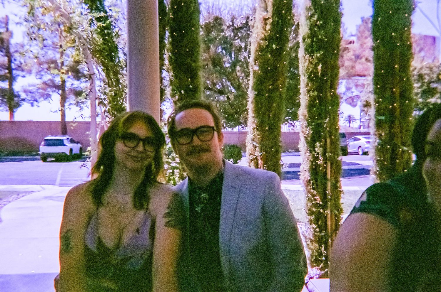 A couple poses together outdoors near ivy-covered pillars, the woman wearing glasses and the man in a gray blazer.