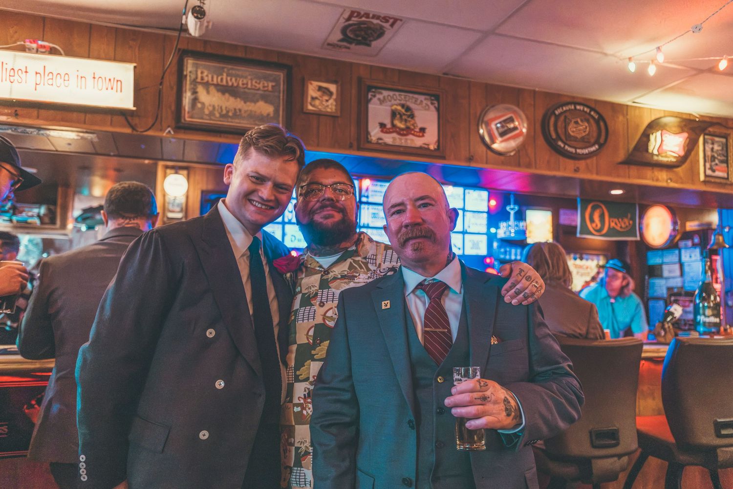 Three men smiling together inside a bar, dressed in suits, with vintage signs and neon lights in the background.