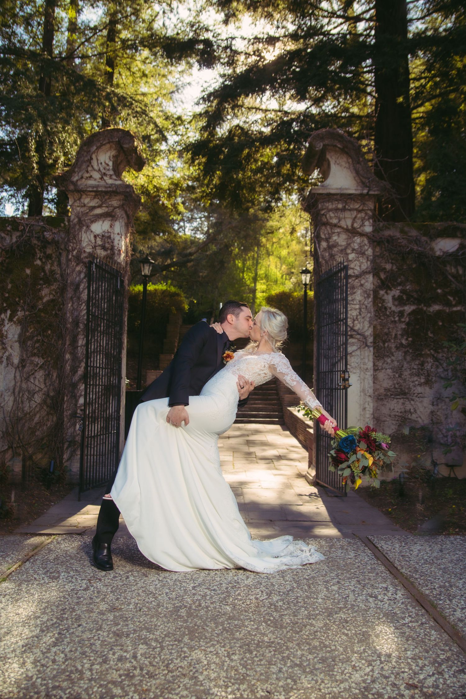 Groom dipping bride for a kiss at ornate garden gates, bride in elegant lace wedding gown with colorful bouquet.