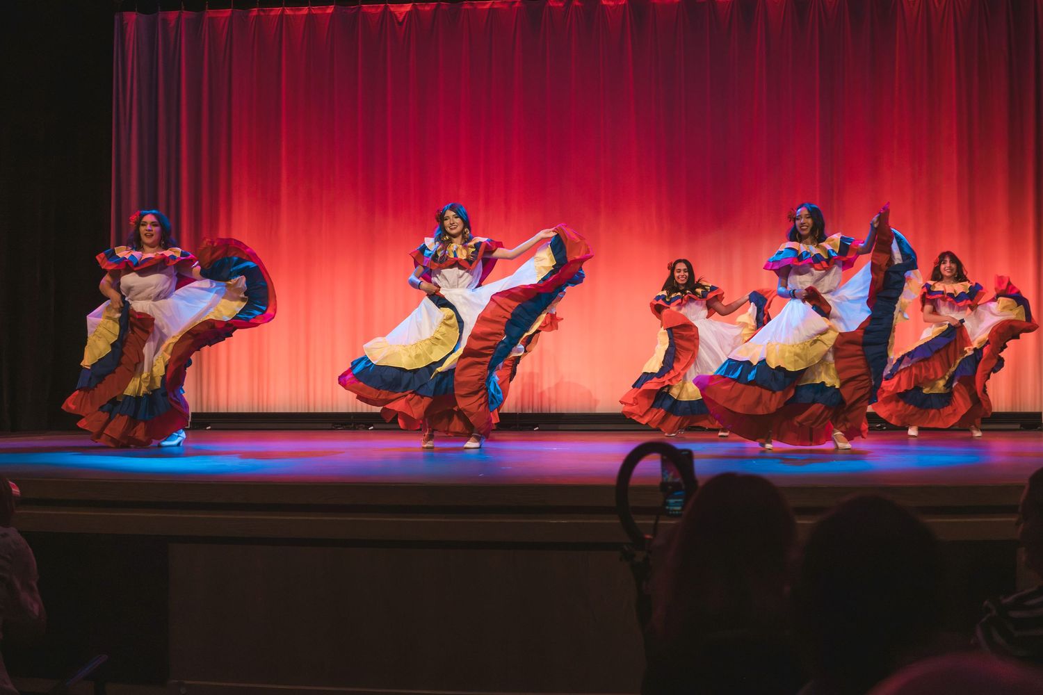 Colorful folkloric dancers in vibrant skirts perform on a red-curtained stage in a lively cultural dance performance.