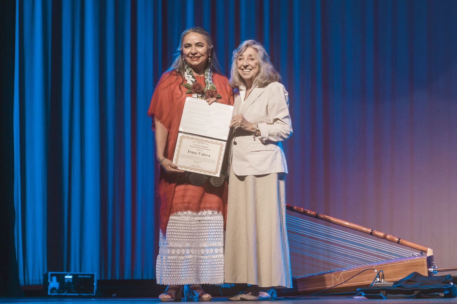 Two women share an award certificate on stage with a dulcimer instrument beside them against a blue curtain.