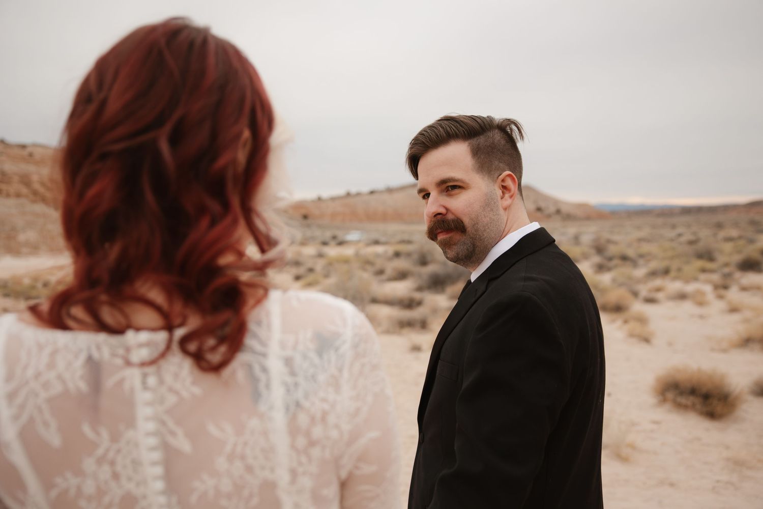 Groom in black tuxedo looks back at red-haired bride in lace dress in an arid desert landscape.