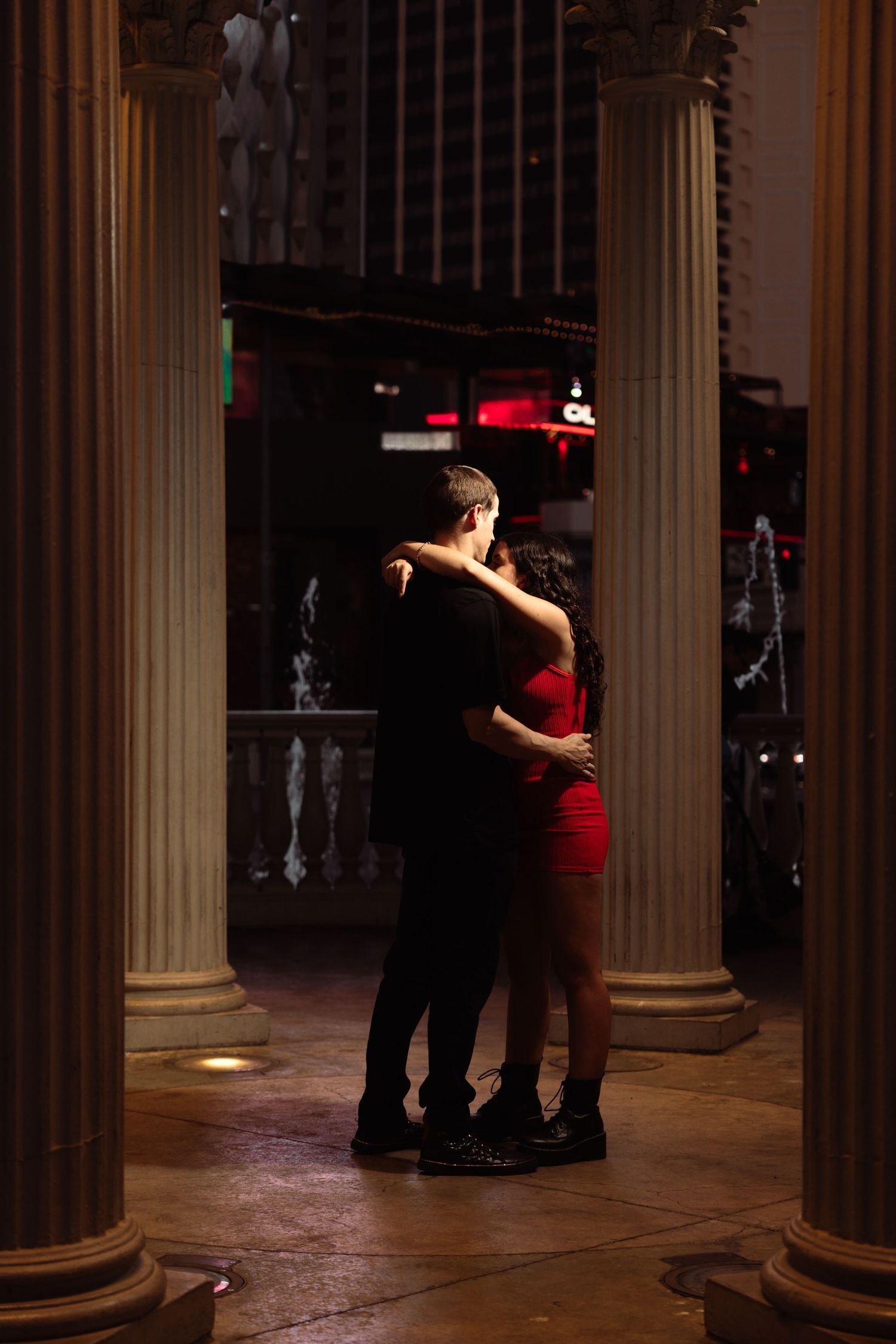 Romantic couple embracing between classical columns at night, woman in red dress and man in black.