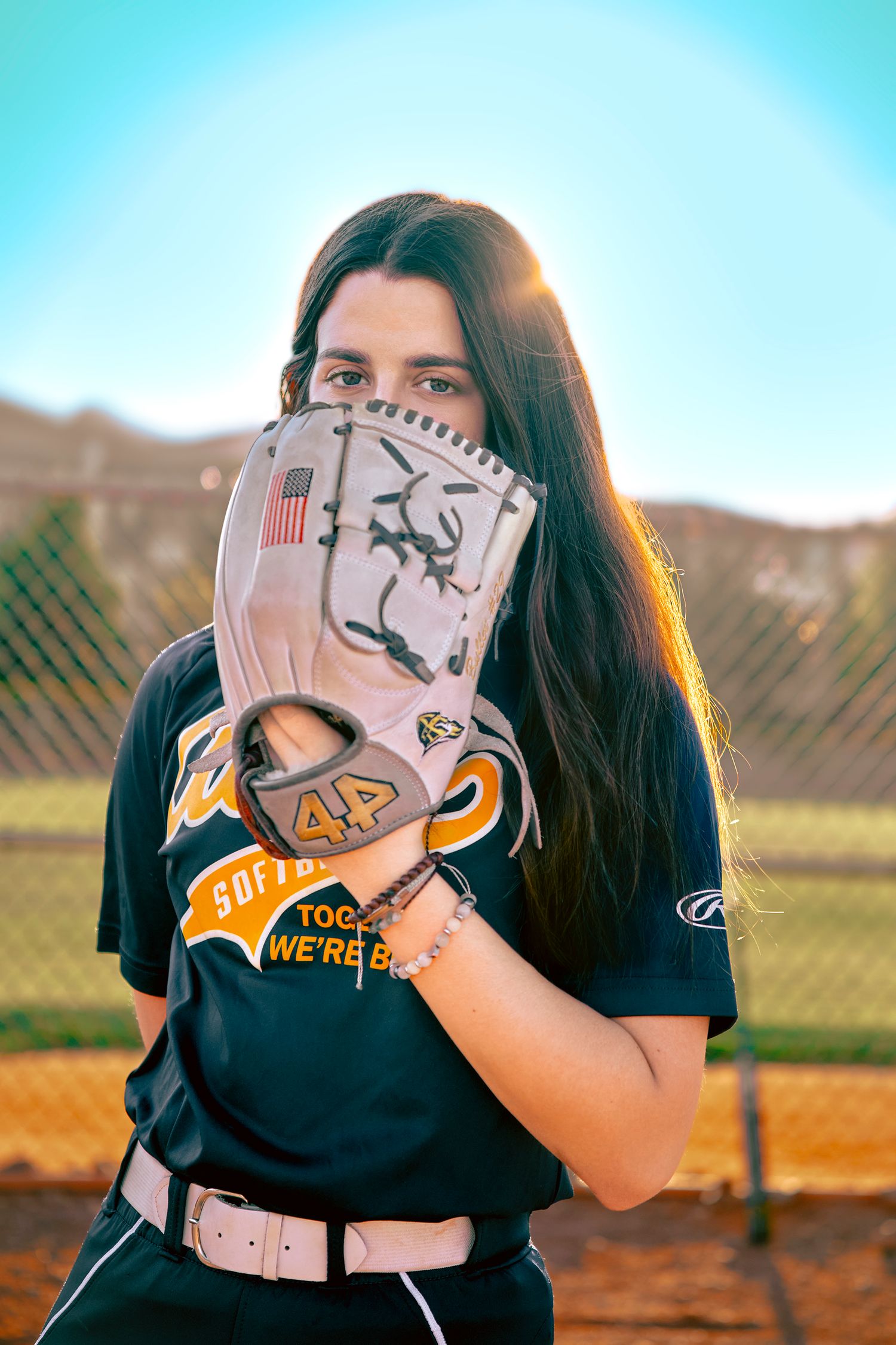 Softball player in jersey holding a Rawlings glove up to her face on a sunny baseball field.