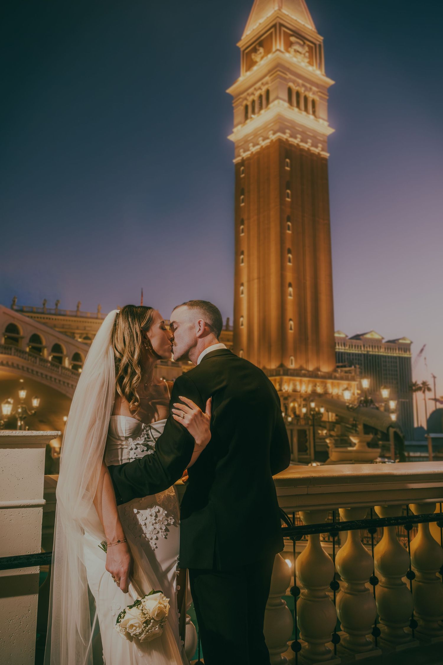 Newlyweds share a passionate kiss with the lit Venetian campanile tower in the Las Vegas night sky.