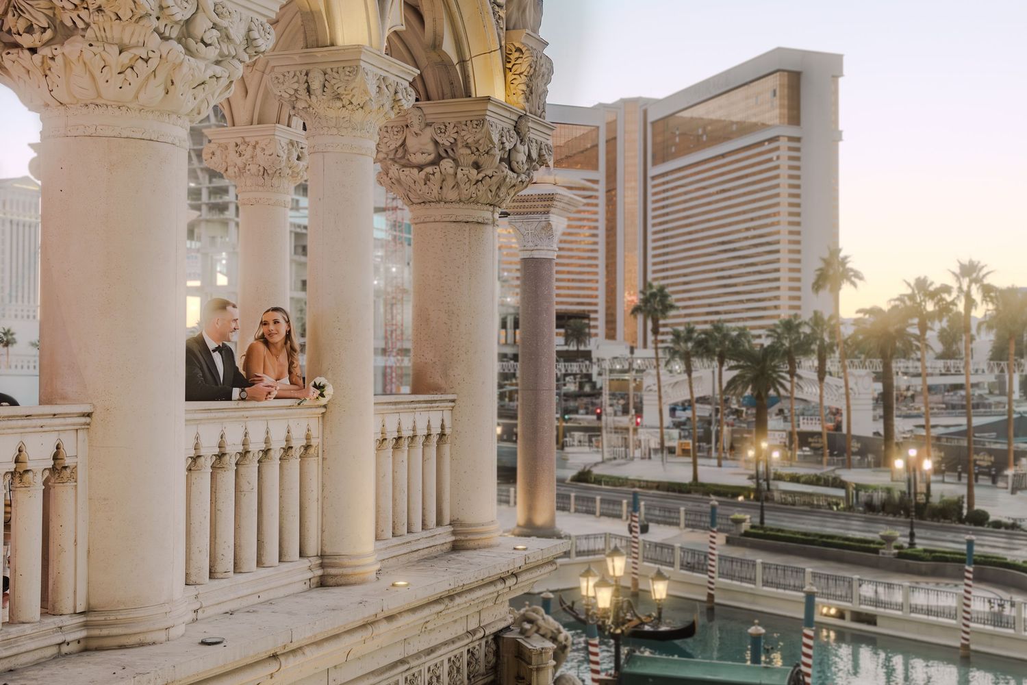 Wedding couple poses on ornate Venetian-style balcony with Las Vegas Strip skyline at sunset.