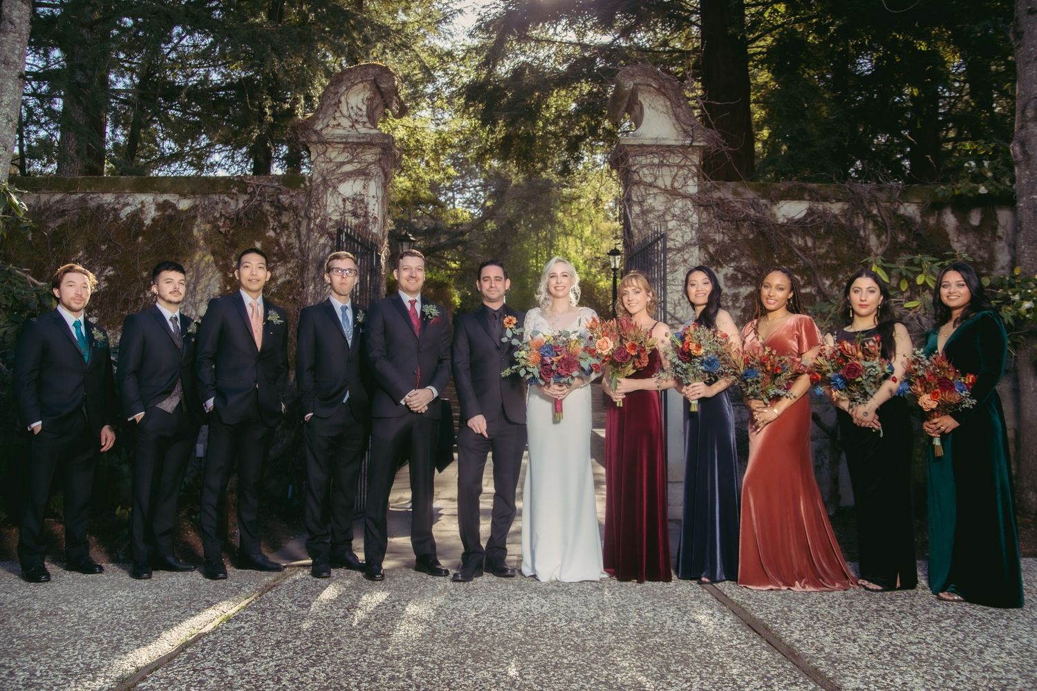 Wedding party poses formally before ornate stone gates, bridesmaids in jewel-tone velvet gowns holding autumn bouquets.