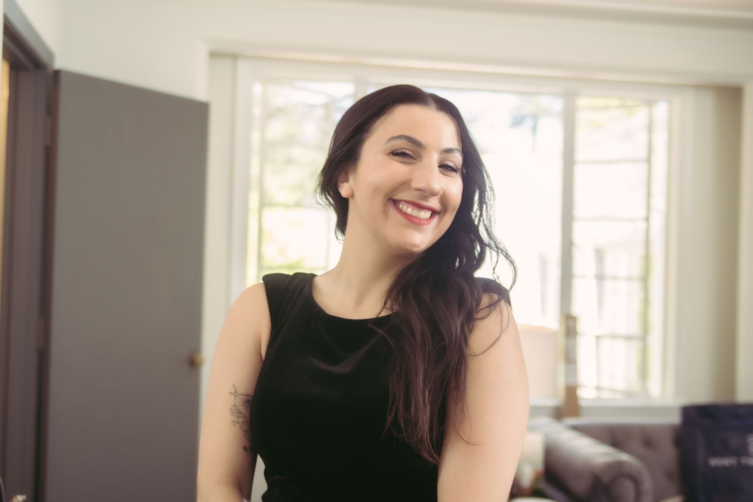Laughing woman with long dark hair in black sleeveless dress in bright room.