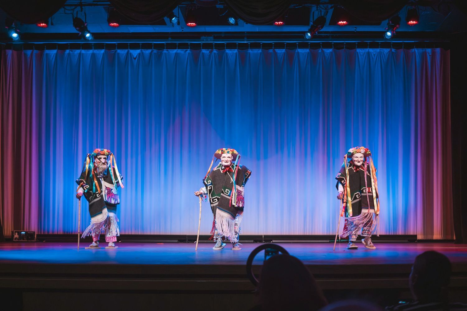 Three performers in colorful traditional costumes and masks dance on a blue-curtained theater stage.