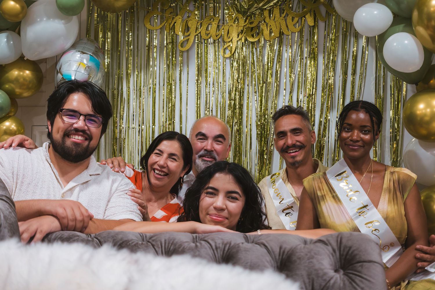 Six friends posing together at a festive party with gold and white balloon decorations and a shimmery backdrop.