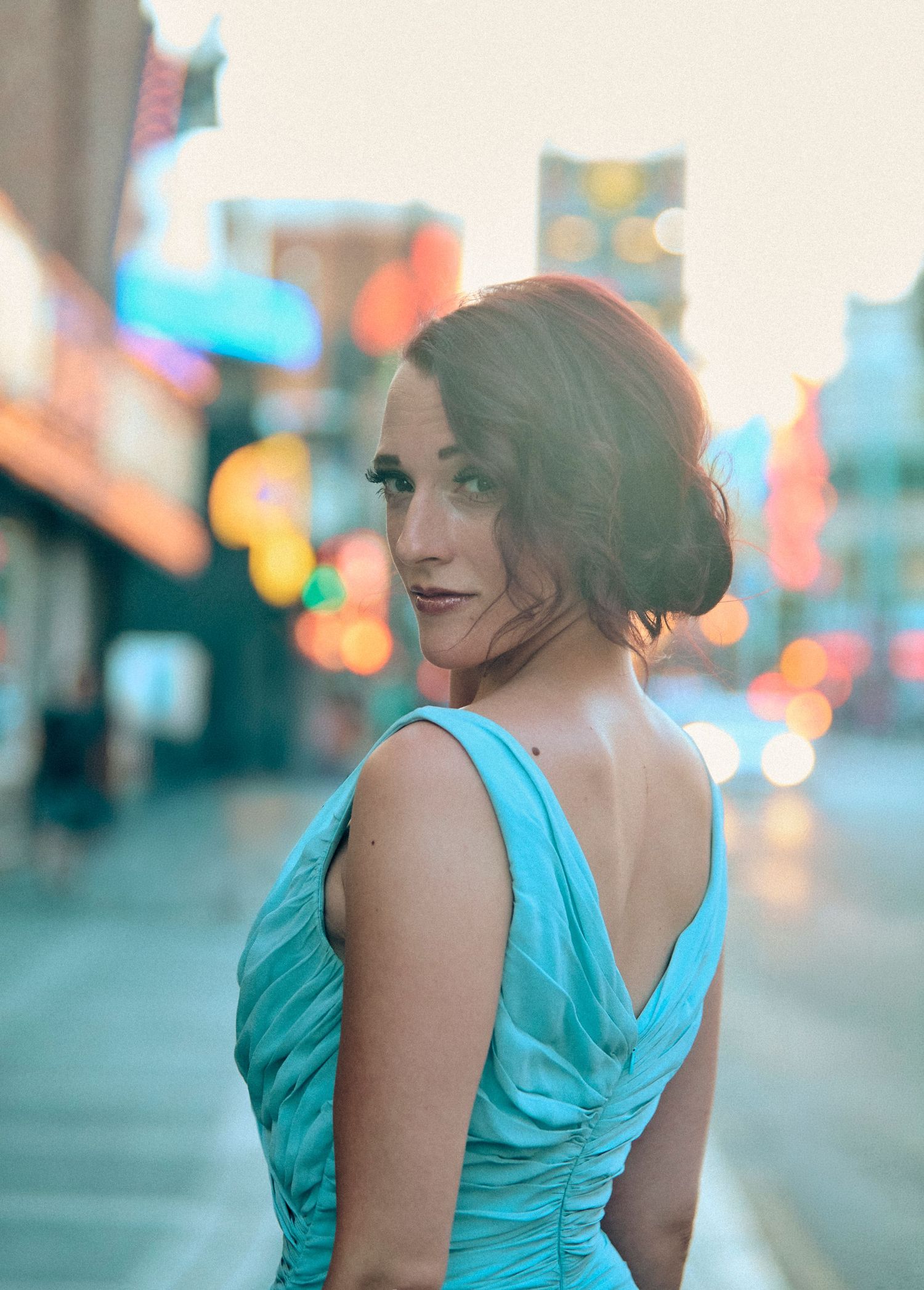 Woman in turquoise backless gown poses on city street at dusk with bokeh lights in background.