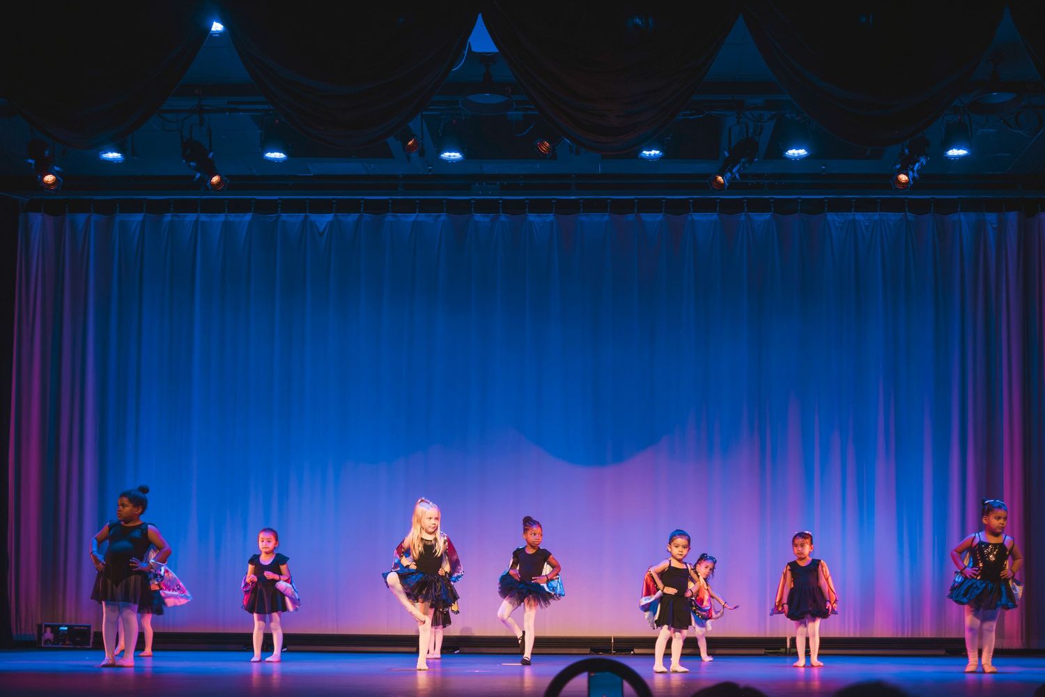 Young children in black costumes perform a dance recital on a blue-lit stage with curtains.