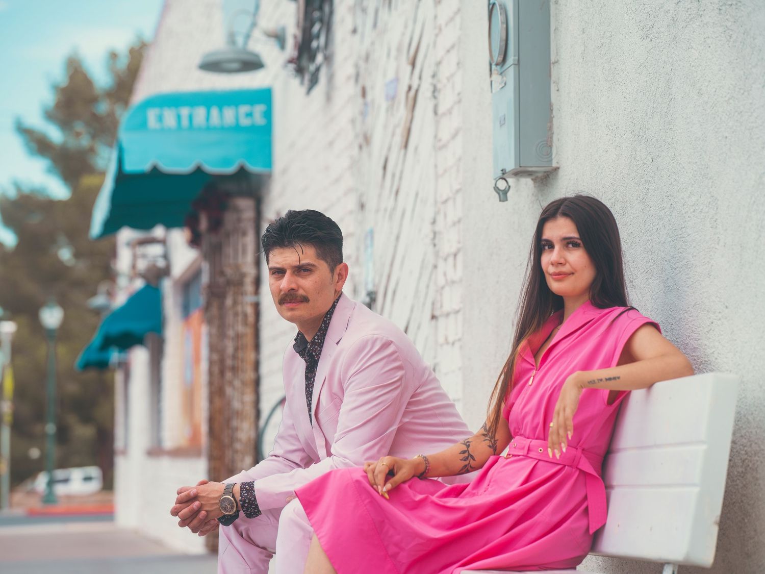 A man in a pink suit and woman in a pink dress pose outside a building with an entrance sign.