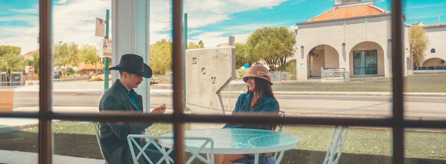 Two people wearing hats sit at a white outdoor table, viewed through a window in a sunny small-town setting.