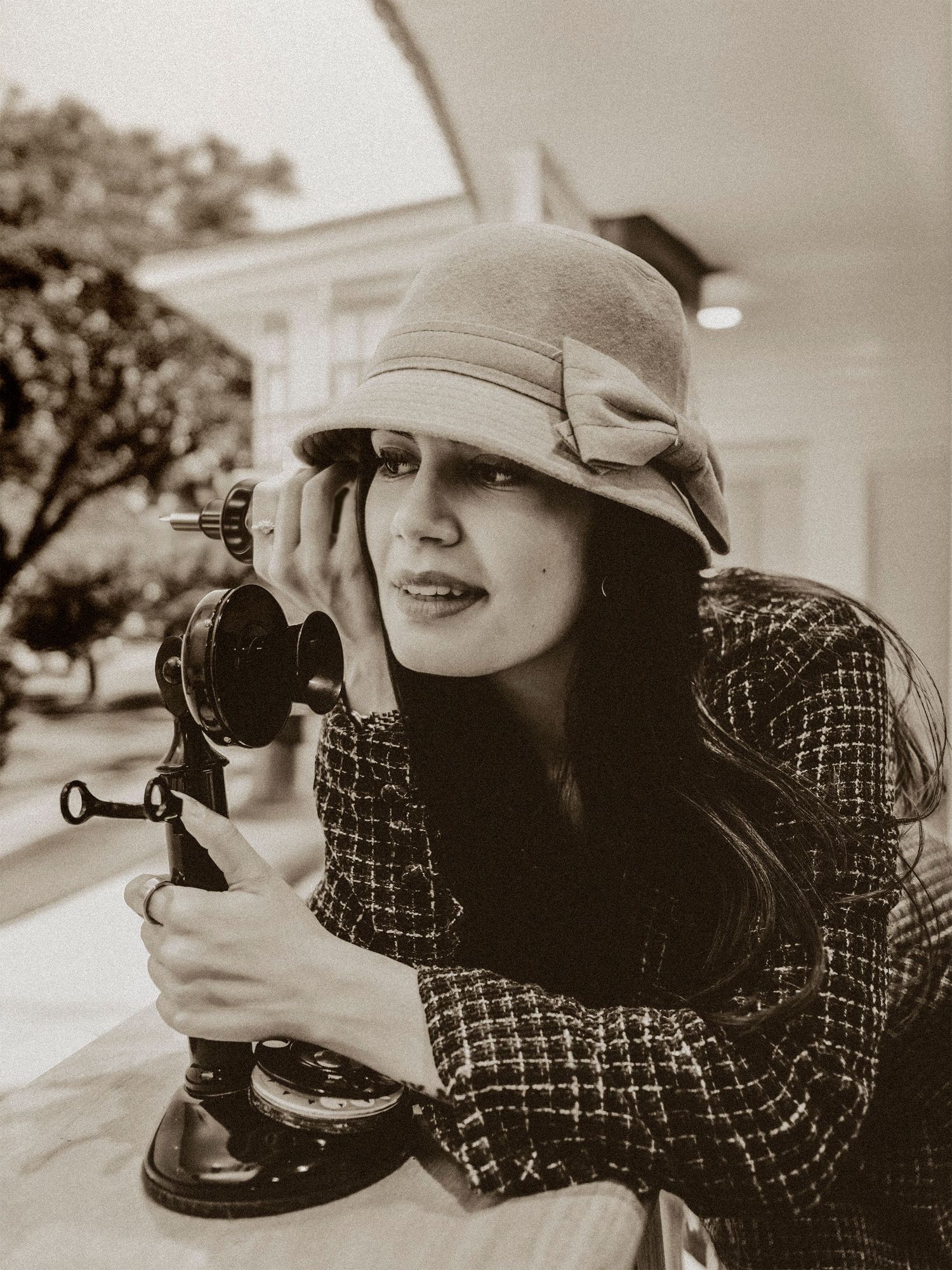 Vintage-style woman in cloche hat and tweed jacket holding antique candlestick telephone outdoors.