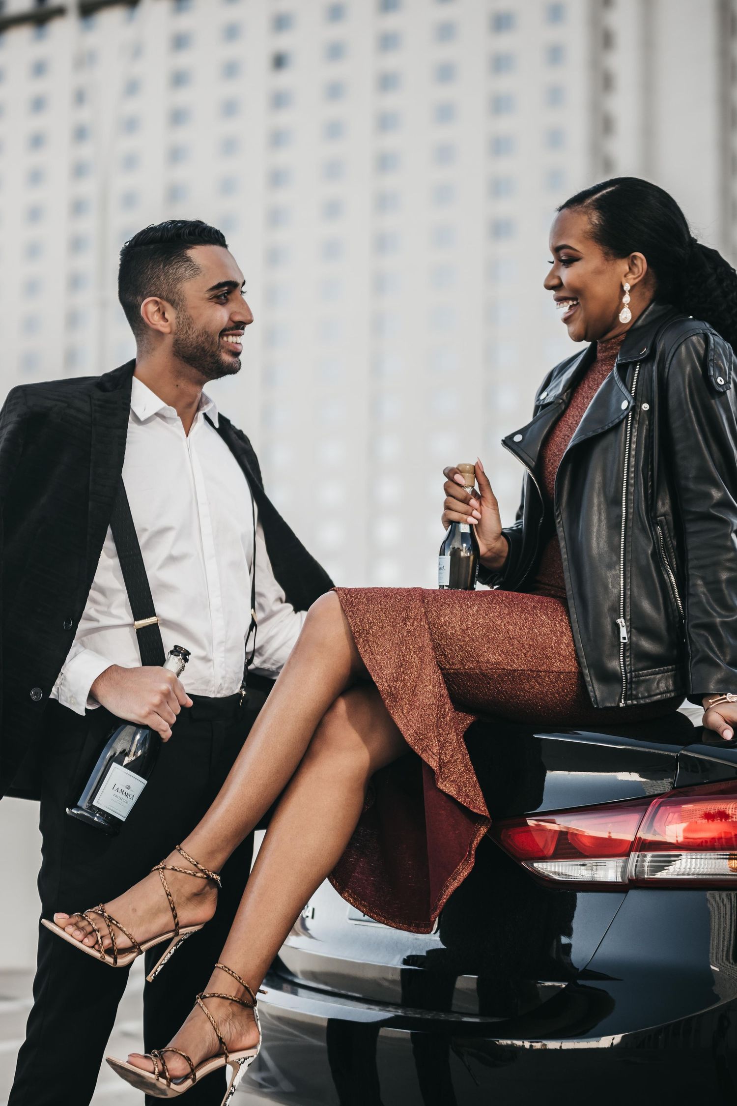 Stylish couple leaning on a luxury car, holding champagne bottles, dressed in elegant attire outdoors.