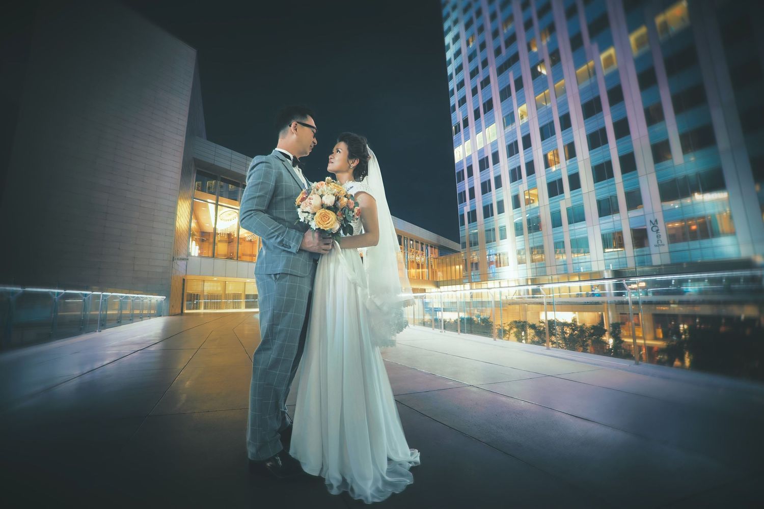 Bride and groom share a romantic moment on a rooftop at night with illuminated city buildings in the background.