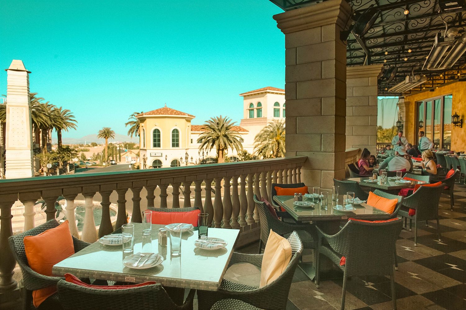 Elegant outdoor restaurant terrace with white tables, palm trees, and Mediterranean architecture under a bright blue sky.