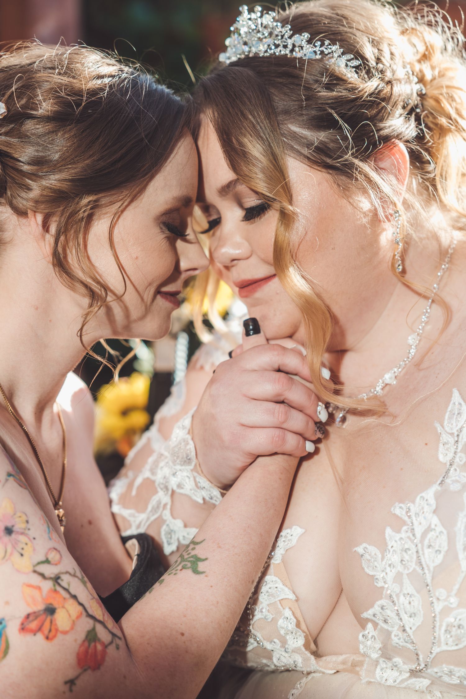 Two brides hold hands and touch foreheads intimately, one wearing crystal tiara and lace wedding gown.