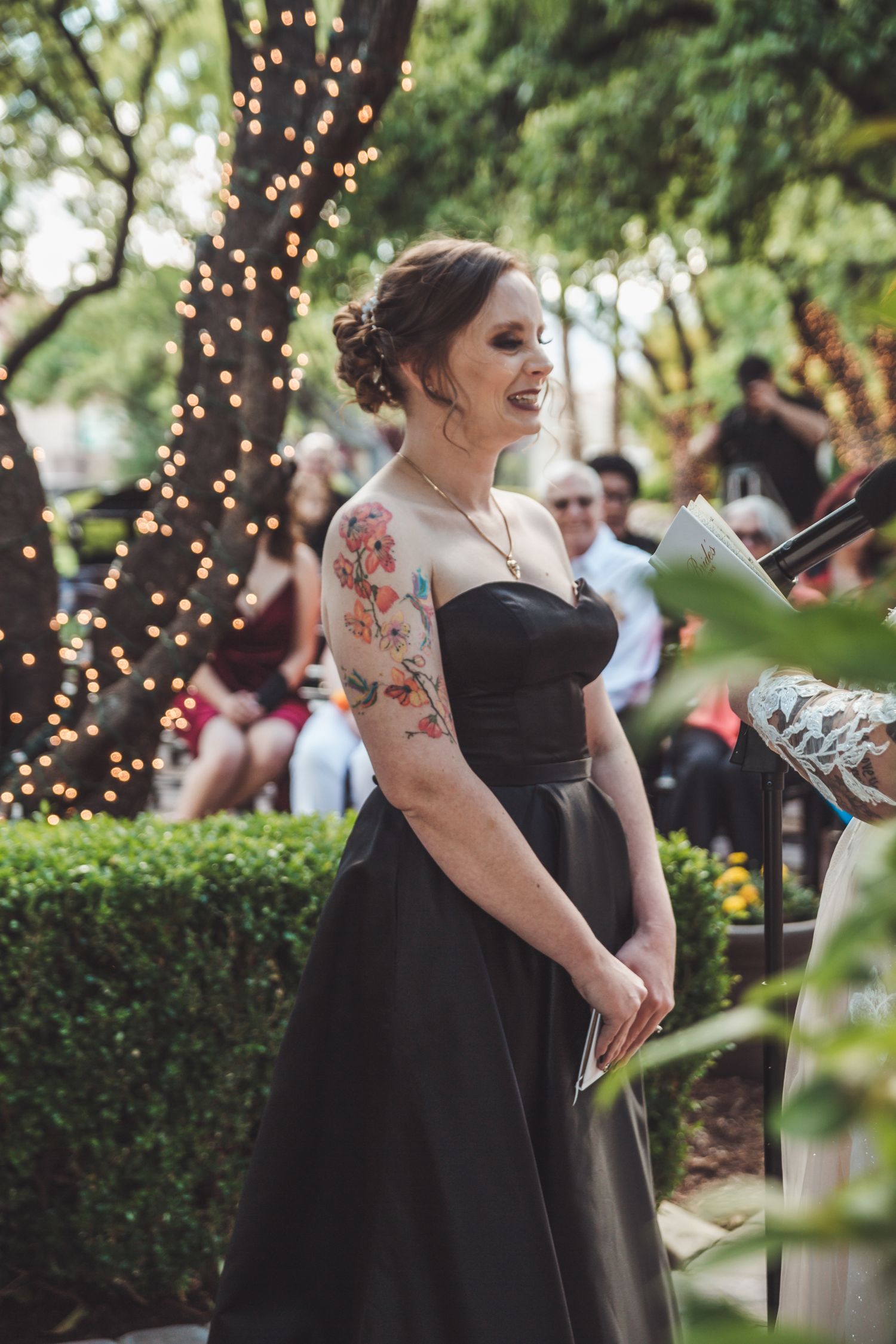 Woman in black strapless gown with floral arm tattoo smiling during outdoor wedding ceremony.