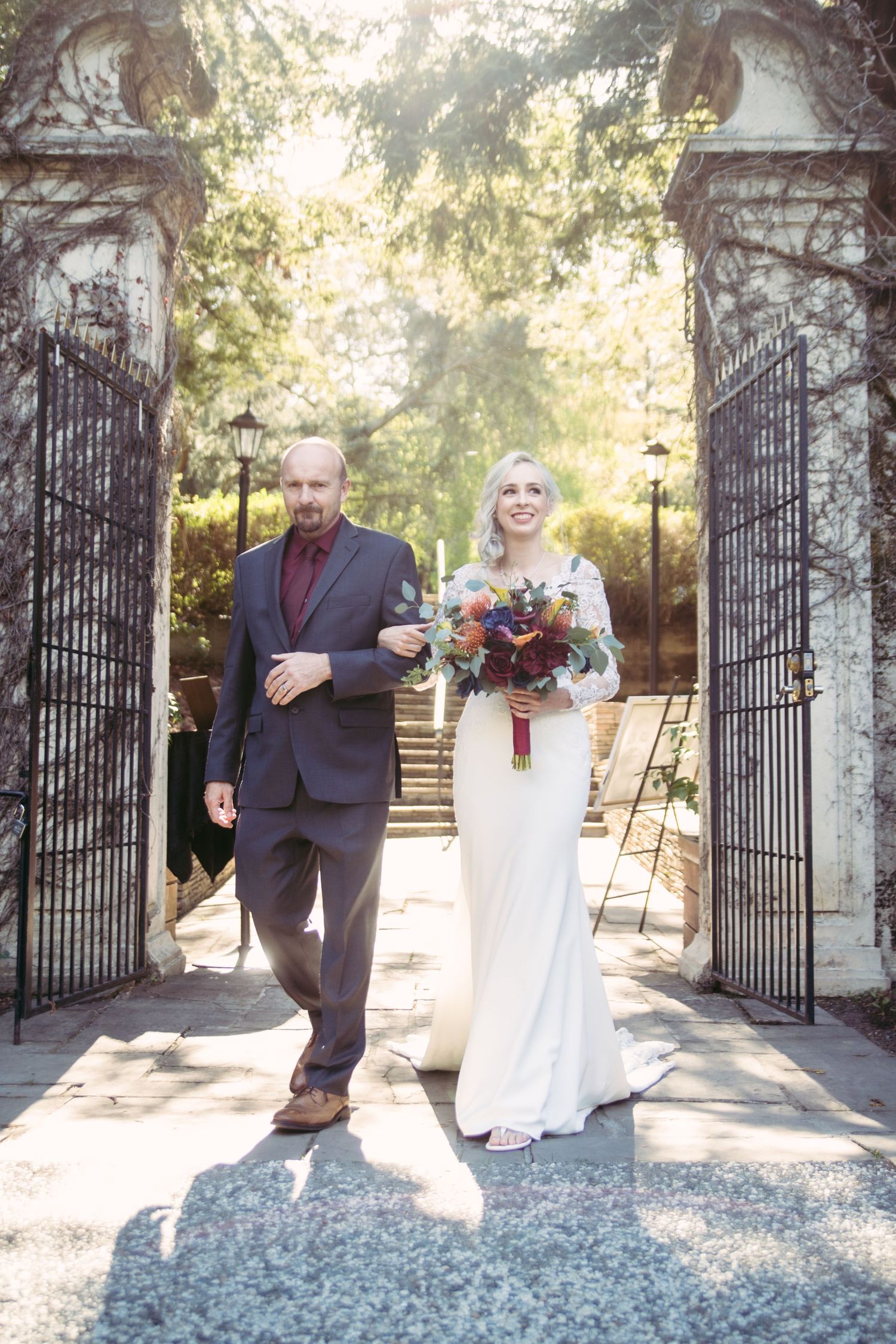 A bride in a white lace gown and her escort emerge through vintage iron gates in golden sunlight at a wedding.
