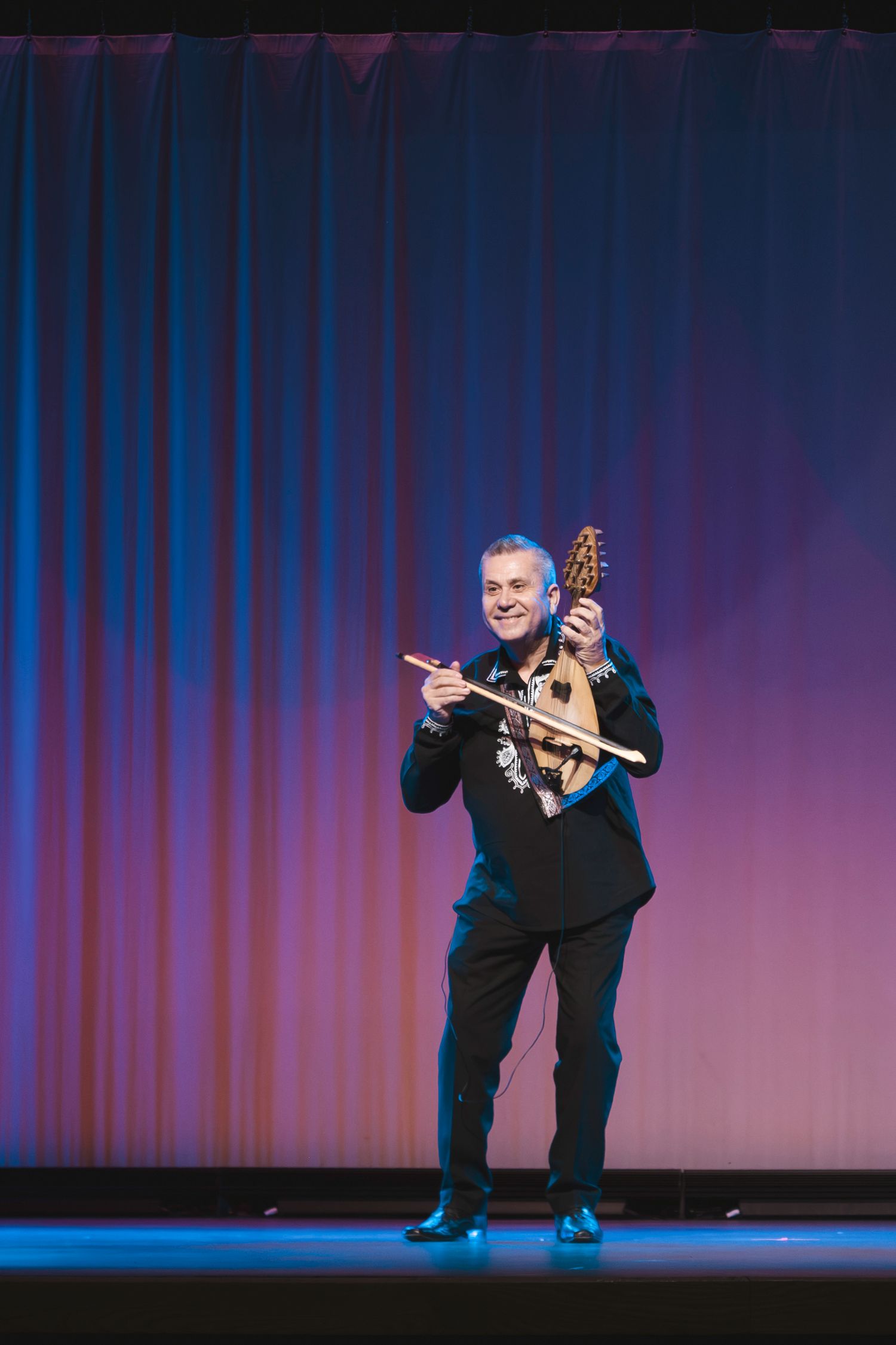 Smiling male musician performing on stage, playing a decorative string instrument in black attire with colorful curtain backdrop.
