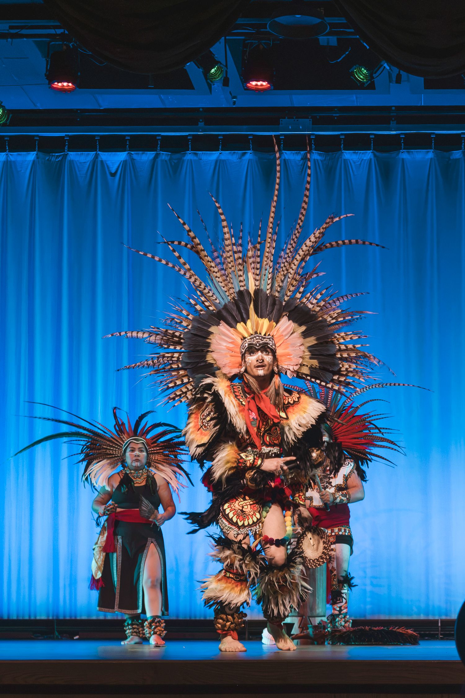 Aztec dancers in traditional feathered regalia perform on a blue-lit stage during a cultural ceremony.