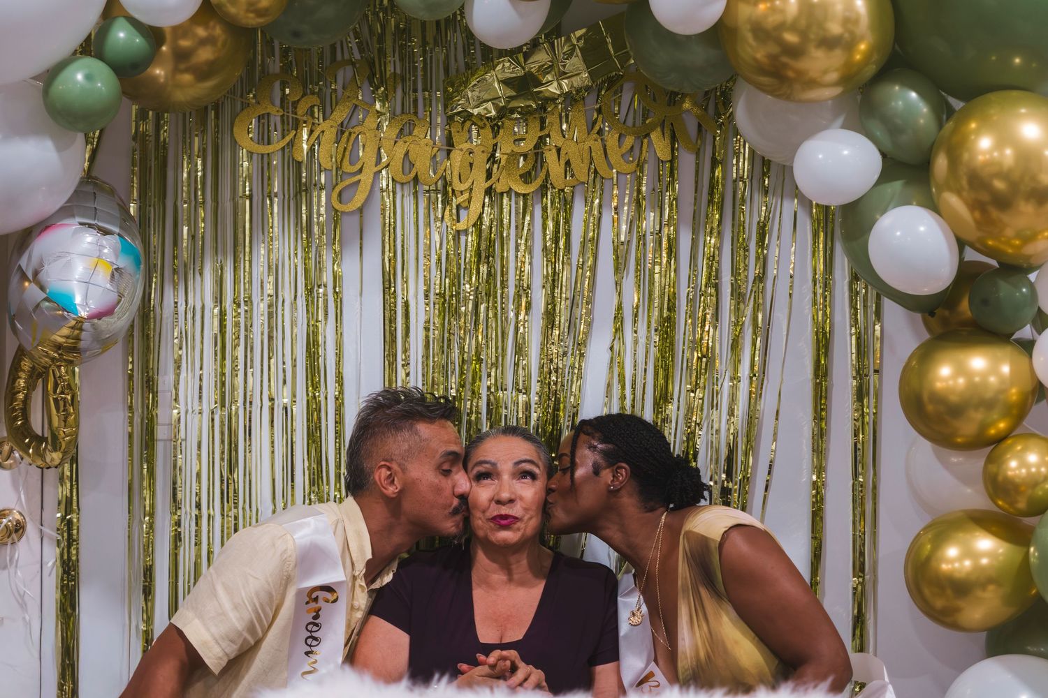 Two people kissing a woman on the cheeks at an engagement party with gold and green balloon decorations.