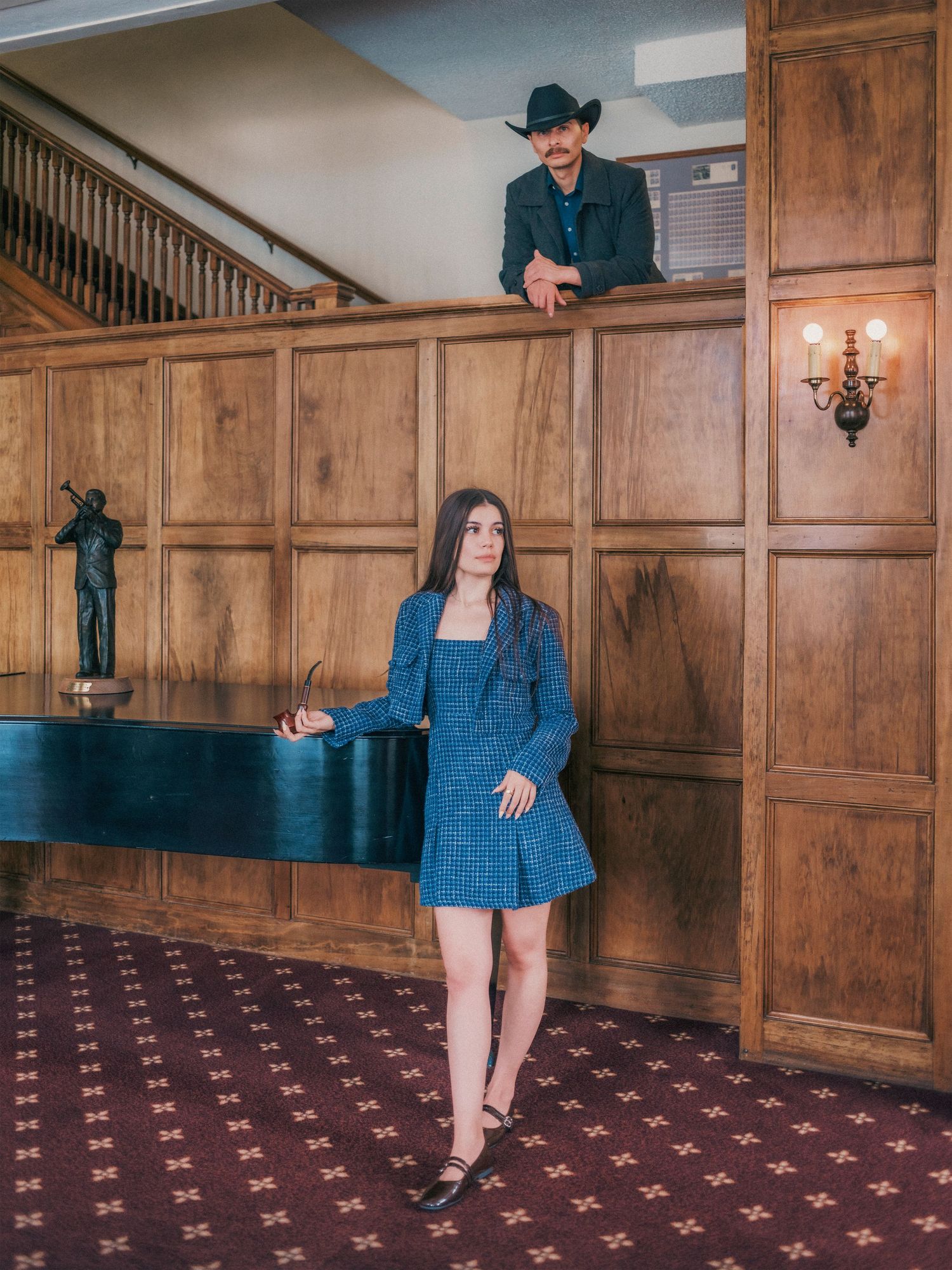 Woman in blue tweed mini dress stands in wood-paneled lobby while man in cowboy hat leans over upper railing.
