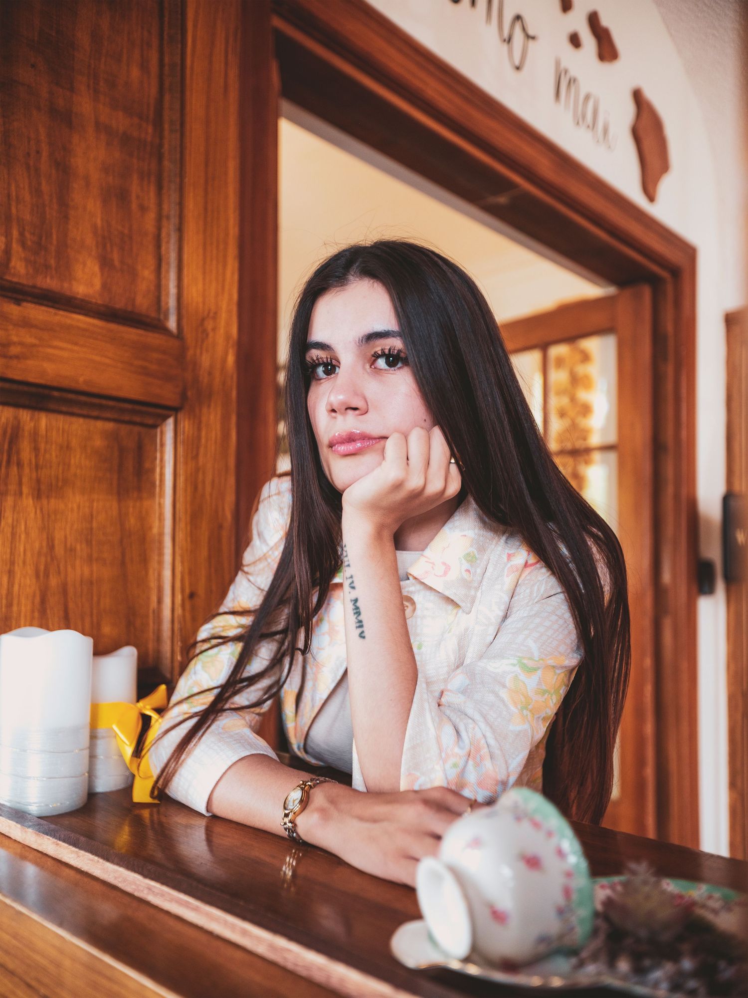 Young woman with long dark hair resting chin on hand at wooden counter with teacup and candles.